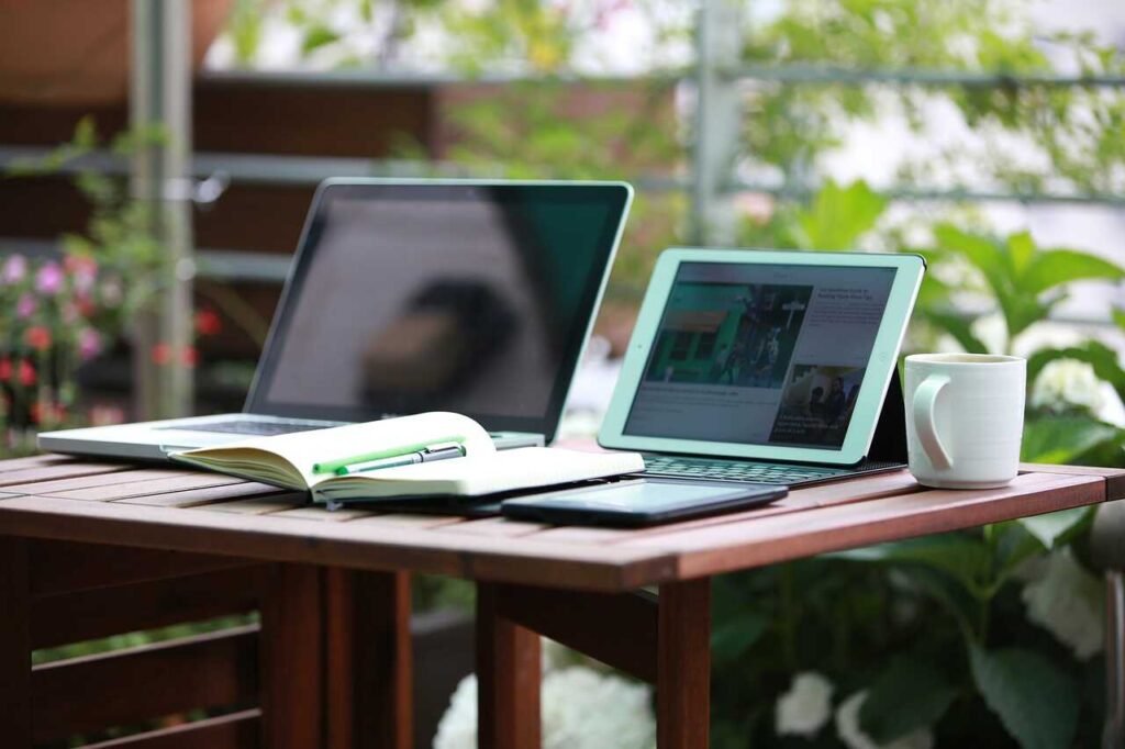 Imagen de una laptop, una tablet, un taza con cafe y una libreta abierta encima de una mesa chocolate de cafeteria